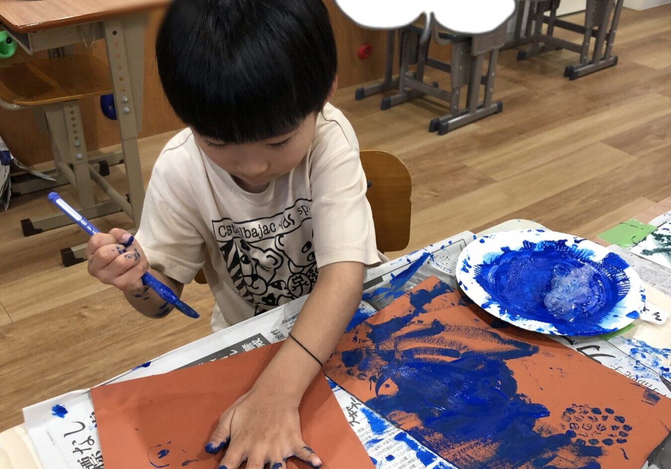 Child in a classroom finger-painting with blue paint on orange paper, hands covered in blue paint; speech bubble reads えいっ！！