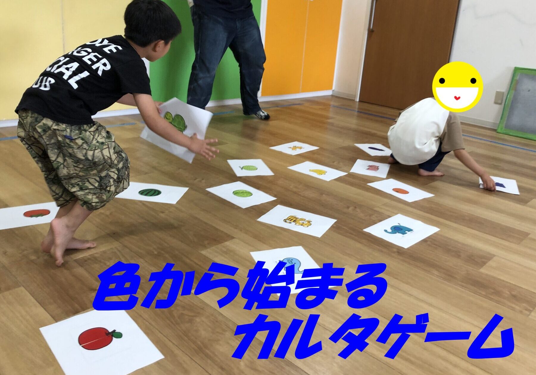 Children playing a floor-based sorting game with printed fruit pictures scattered on white sheets in a brightly colored room. One child is reaching for a card while another sits with a big emoji covering their face.