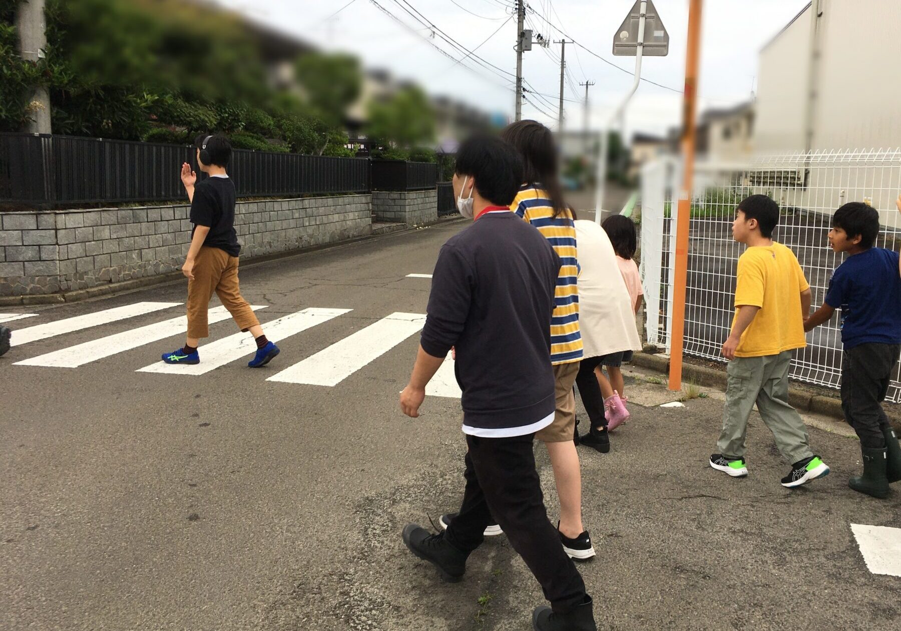 Group of kids walking across a zebra crosswalk on a residential street, with a boy in a black shirt waving as they walk past a fence and power lines.