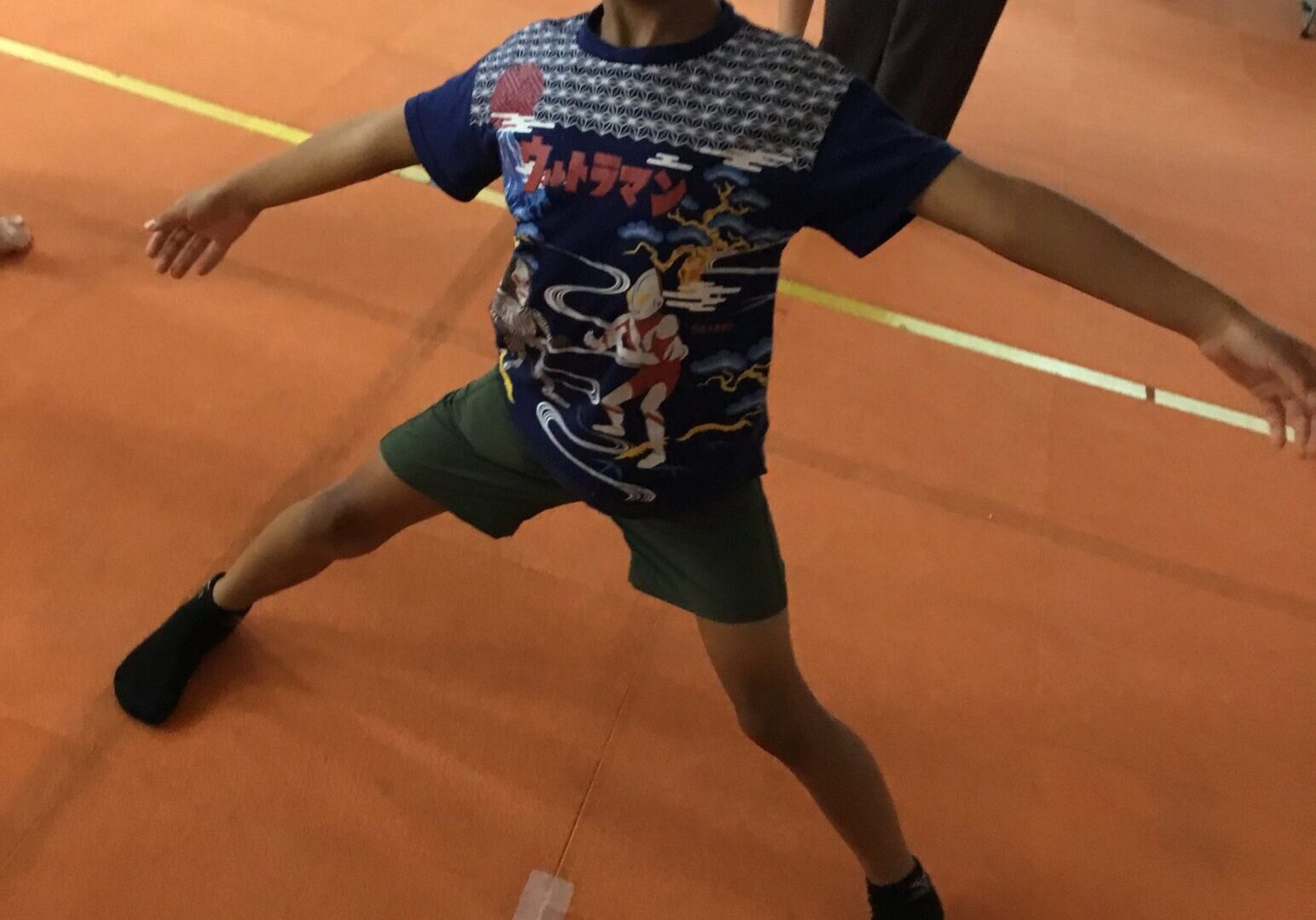 Boy in a blue graphic T-shirt and green shorts balancing with arms outstretched in a gymnasium on an orange floor.